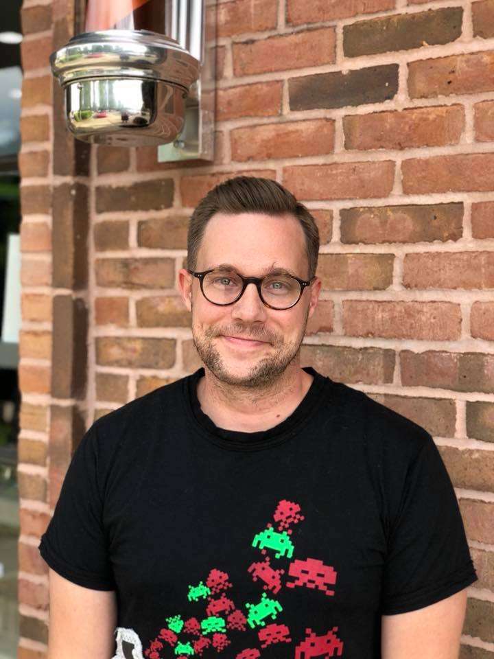 Man with glasses smiling in front of a brick wall, wearing a black t-shirt with a colorful pixelated design. - K. Charles & Co. in Schertz, TX