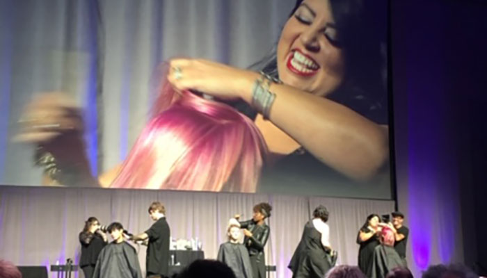 Hair stylists demonstrate hairstyling techniques on models at an event, with a large screen displaying a close-up of a smiling woman having her hair styled. - K. Charles & Co. in Schertz, TX