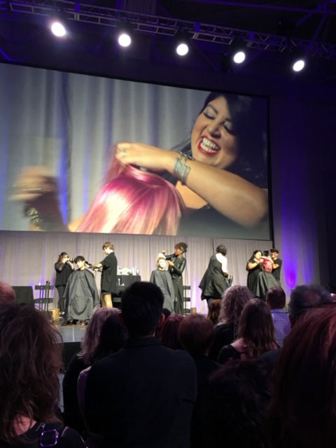 A hairstyling demonstration on stage at a convention, with a large screen displaying a smiling woman holding colorful hair extensions. audience members watch attentively. - K. Charles & Co. in Schertz, TX