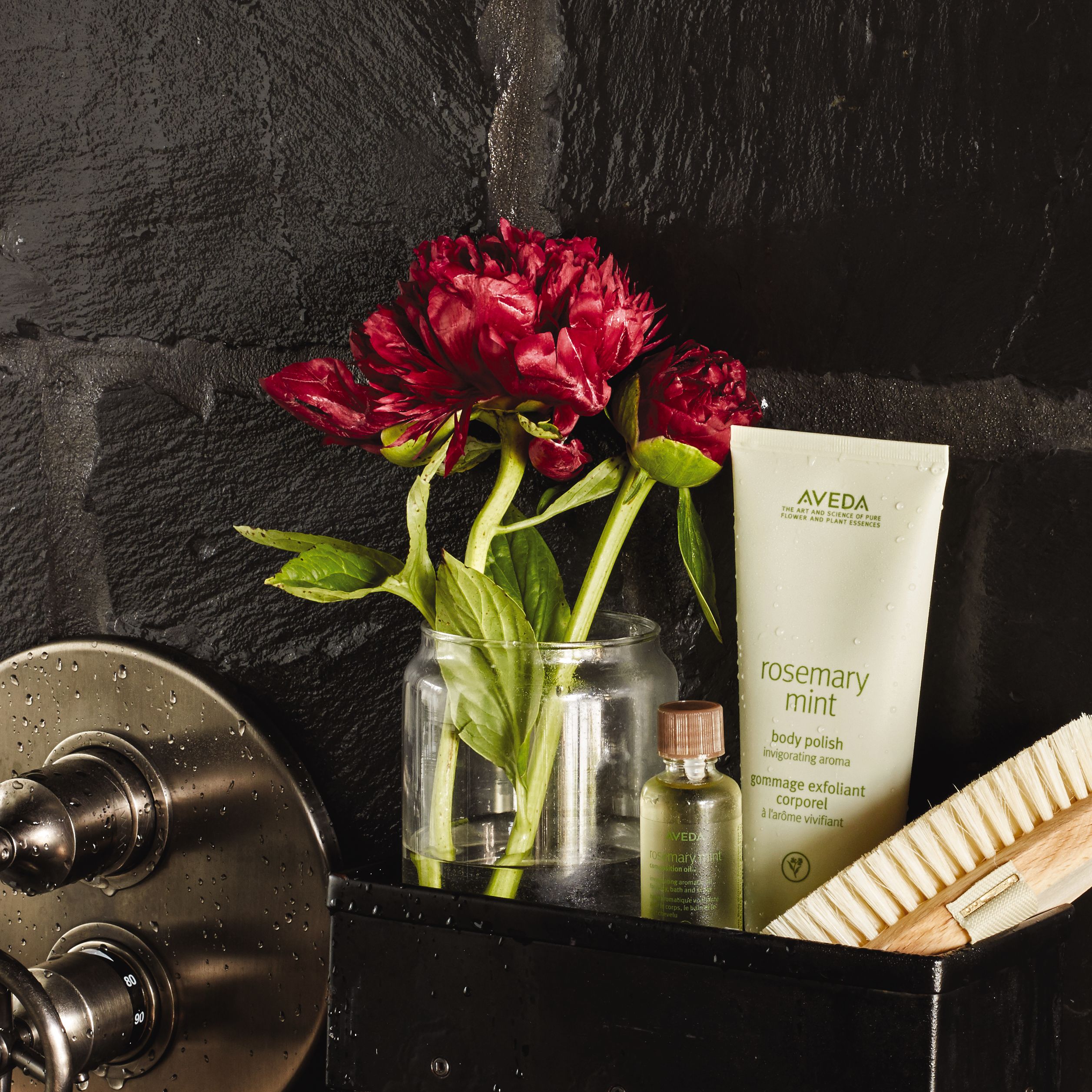 Glass jar with lush red peonies next to aveda body polish and a wooden brush on a black tiled surface. - K. Charles & Co. in Schertz, TX