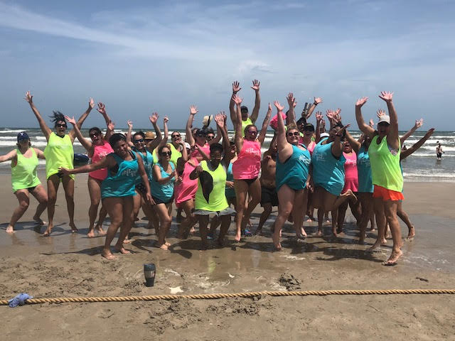 A group of people with styled hair in colorful beachwear cheerfully posing with raised arms on a sandy beach, with waves in the background. - K. Charles & Co. in Schertz, TX