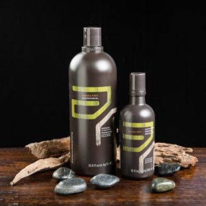 Two bottles of men's grooming products on a wooden surface, one large and one small, with gray and yellow labeling, surrounded by rocks and wood pieces. - K. Charles & Co. in Schertz, TX