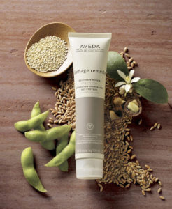 Aveda damage remedy hair care product on a wooden surface surrounded by a bowl of seeds, loose grains, and green pods. - K. Charles & Co. in Schertz, TX
