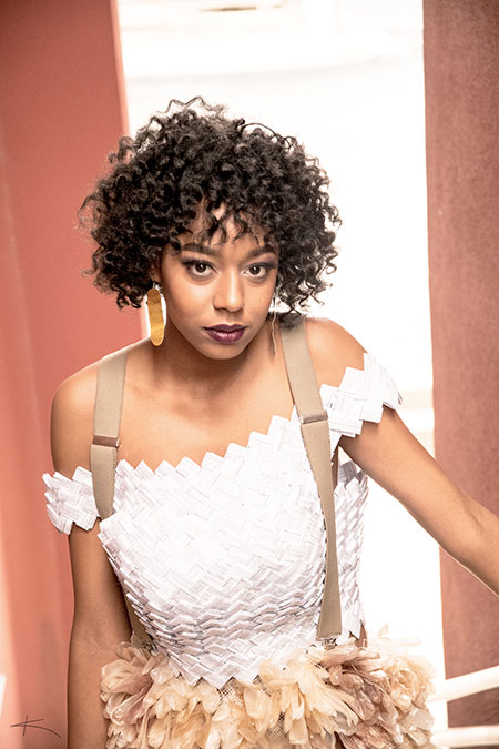 A woman with curly hair wearing a creative dress with white top and beige ruffled skirt, looking intently at the camera in a red-hued room. - K. Charles & Co. in Schertz, TX