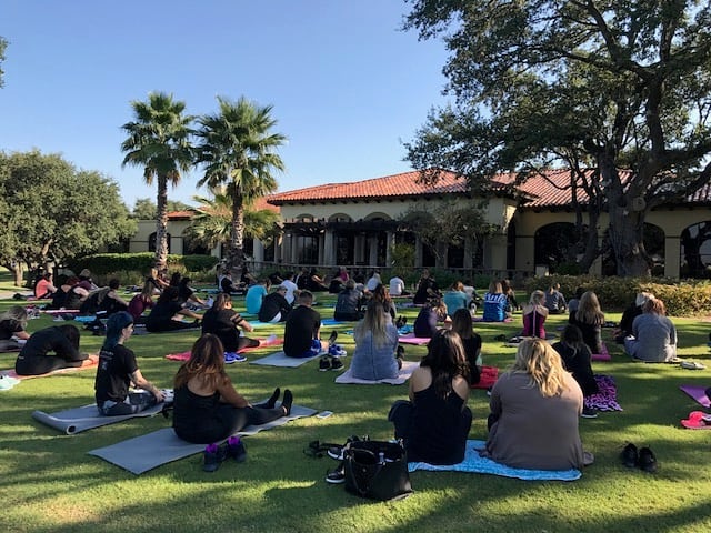 Outdoor yoga class with participants seated on mats in a park, facing an instructor, with trees and a building in the background. - K. Charles & Co. in Schertz, TX