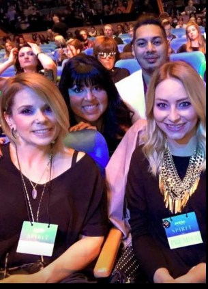 Four people sitting in a theater, three women and one man, smiling at the camera, wearing event lanyards. - K. Charles & Co. in Schertz, TX