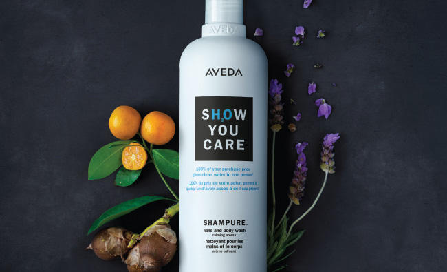 Aveda shampoo bottle with the text "show you care" surrounded by orange fruits, a leaf, and scattered purple flowers on a dark background, perfect for any salon. - K. Charles & Co. in Schertz, TX