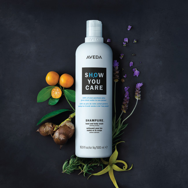 Aveda shampoo bottle with the text "show you care" surrounded by purple flowers, citrus fruits, and green leaves on a dark background. - K. Charles & Co. in Schertz, TX