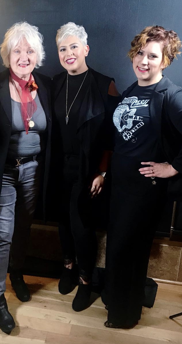 Three women smiling for a photo, standing in a room with wooden flooring and blue walls. they have diverse hair colors and styles, wearing casual to stylish outfits. - K. Charles & Co. in Schertz, TX