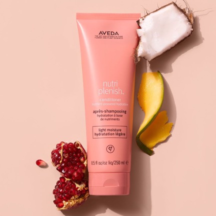 Aveda nutriplenish conditioner bottle alongside ingredients: coconut half, mango slices, and pomegranate on a beige background, ideal for maintaining your salon hairstyle. - K. Charles & Co. in Schertz, TX