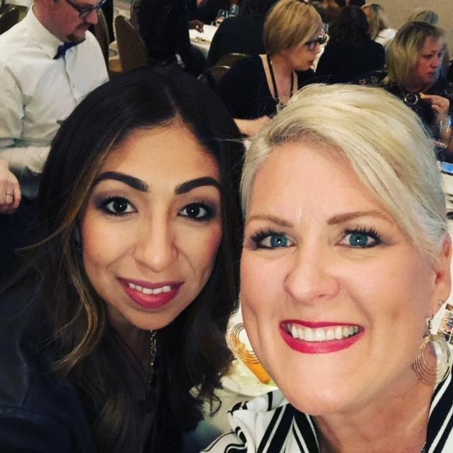 Two women smiling for a selfie at a crowded event, one with light hair and one with dark hair, both dressed in evening wear. - K. Charles & Co. in Schertz, TX