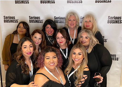 Group of ten smiling women posing for a photo at a "serious business" conference, standing in front of a branded backdrop. - K. Charles & Co. in Schertz, TX
