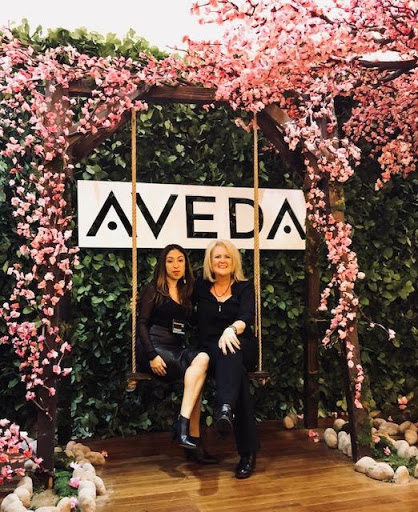 Two women sitting on a swing under a floral arch with the sign "aveda" at a promotional event. - K. Charles & Co. in Schertz, TX