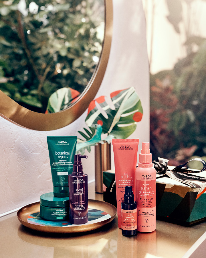 A collection of aveda beauty products arranged on a tray before a round mirror, with lush greenery in the background. - K. Charles & Co. in Schertz, TX