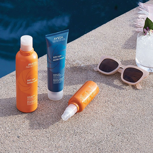 Three Aveda sun care products arranged beside sunglasses and a flower by a swimming pool, ideal for maintaining hair color protection in the sun. - K. Charles & Co. in Schertz, TX