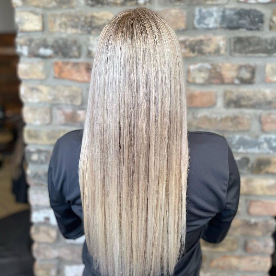A person with long, straight blonde hair colored at a salon, standing in front of a stone wall, viewed from behind. - K. Charles & Co. in Schertz, TX