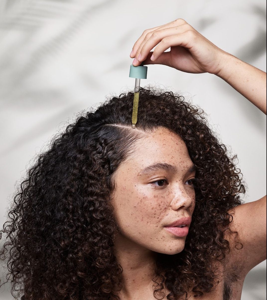 A woman with curly hair applies oil to her scalp using a dropper. - K. Charles & Co. in Schertz, TX