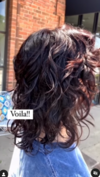 Back view of a person with a salon-styled shoulder-length curly hairstyle highlighted with red tones, standing outside a brick building, with a caption that reads "voila!!". - K. Charles & Co. in Schertz, TX