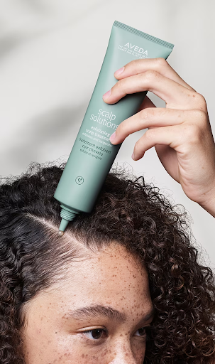 A person applying aveda scalp solutions serum to their scalp from a green tube, focusing on a section of curly hair. - K. Charles & Co. in Schertz, TX