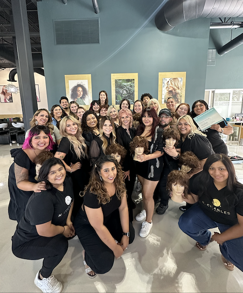 Group of hairstylists in black uniforms posing with mannequin heads at a salon, smiling at the camera. - K. Charles & Co. in Schertz, TX