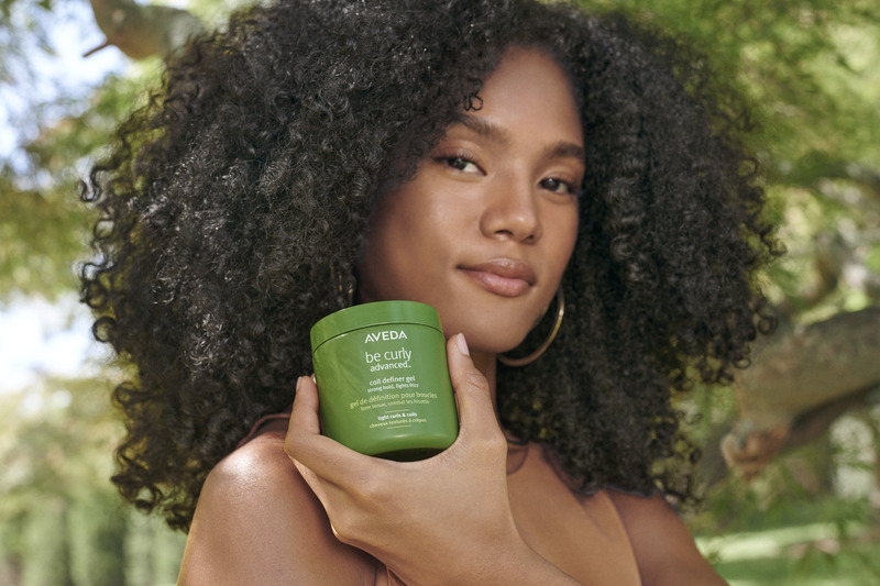 A young woman with curly hair holding a jar of aveda be curly hair product, standing outdoors with greenery in the background. - K. Charles & Co. in Schertz, TX