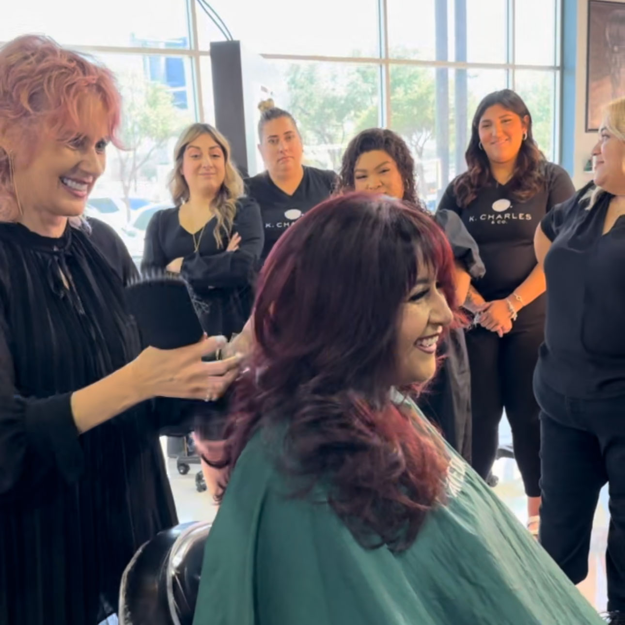 A group of hairstylists watch as a woman with red hair gets her hair styled by another smiling hairstylist in a salon. - K. Charles & Co. in Schertz, TX
