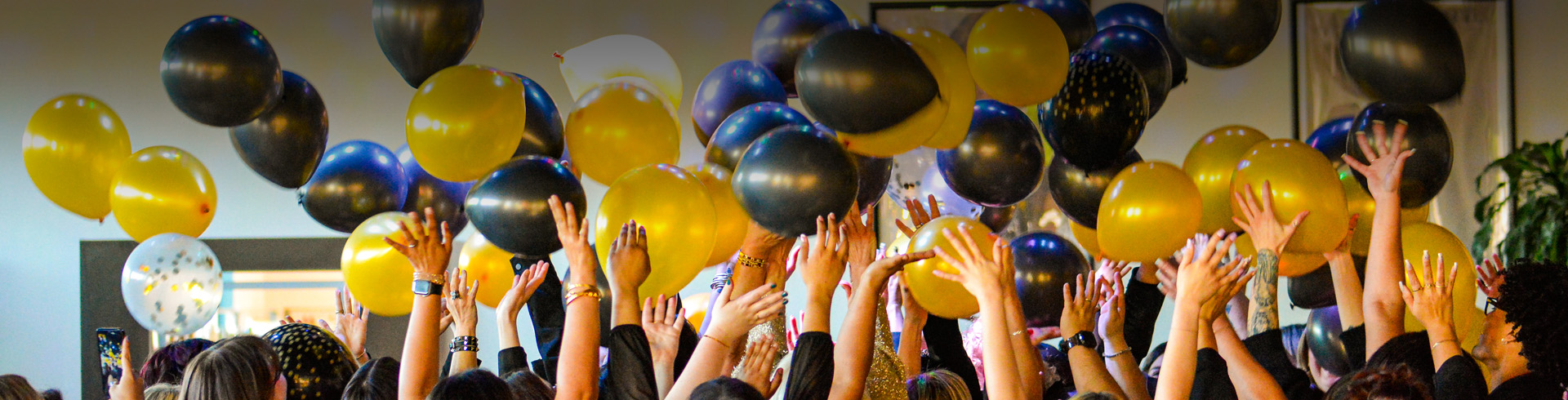 A crowd of people raise their hands towards black and gold balloons floating in the air at an indoor event. - K. Charles & Co. in Schertz, TX