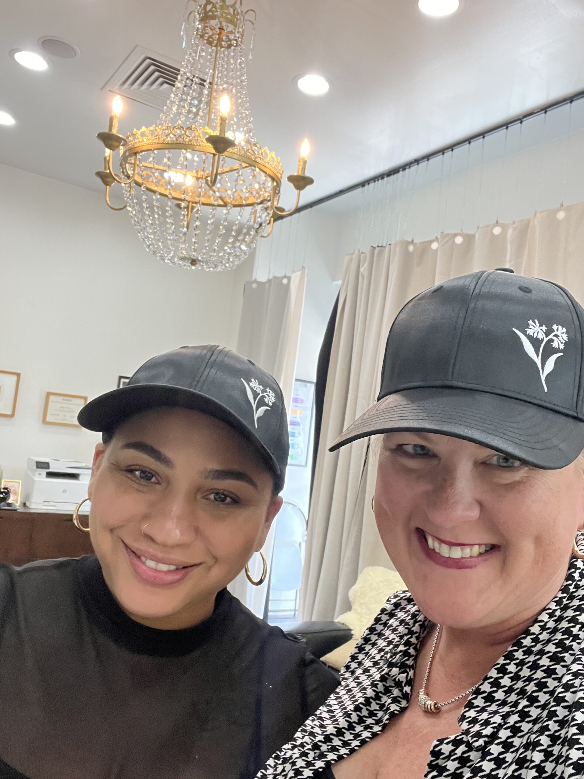 Two women wearing black hats with a white floral logo smile indoors in a well-lit room with a chandelier and framed certificates on the wall. - K. Charles & Co. in Schertz, TX