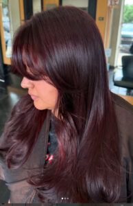 A person with long, straight, dark burgundy hair and layered bangs sitting in a salon chair. - K. Charles & Co. in Schertz, TX
