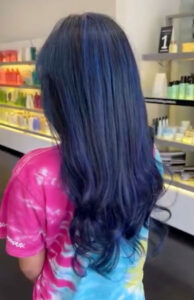 Person with long, wavy, dark blue hair stands in a store, wearing a pink and blue tie-dye shirt. Shelves with colorful bottles line the background. - K. Charles & Co. in Schertz, TX