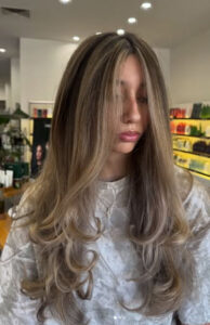 A person with long, layered, wavy ash-blonde hair is standing in a brightly lit salon, wearing a light-colored top. - K. Charles & Co. in Schertz, TX