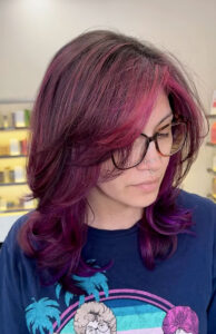 Person with shoulder-length, layered magenta and purple hair, wearing glasses and a blue T-shirt, looks downward in a brightly lit room. - K. Charles & Co. in Schertz, TX