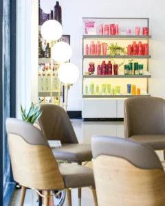 A salon waiting area with beige chairs, a small table, three globe floor lamps, and shelves displaying assorted hair products arranged by color. - K. Charles & Co. in Schertz, TX