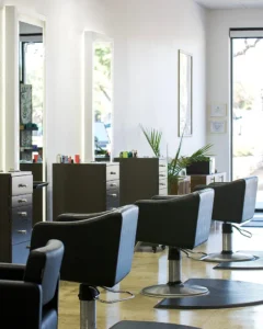 Three modern black salon chairs and styling stations with mirrors in a brightly lit hair salon, featuring minimal decor and large windows. - K. Charles & Co. in Schertz, TX