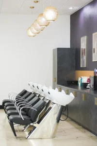 A row of black salon chairs with white washbasins, pendant lights above, and shelves with hair products and equipment along the wall. - K. Charles & Co. in Schertz, TX