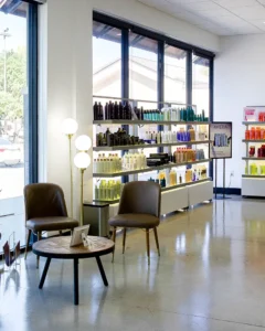 A modern salon waiting area with two brown chairs, a round wooden table, and shelves displaying various hair products by large windows. - K. Charles & Co. in Schertz, TX