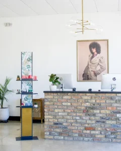 Modern reception area with a brick front desk, two desktop computers, potted plants, product display stand, and a large fashion portrait on the wall. - K. Charles & Co. in Schertz, TX