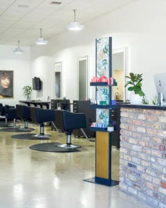 Modern hair salon interior with several black styling chairs, mirrors, a brick reception desk, display shelves with products, potted plants, and wall art. - K. Charles & Co. in Schertz, TX