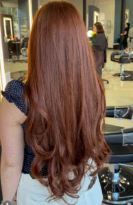 Person with long, wavy auburn hair stands in a modern salon, facing away from the camera. Salon chairs, mirrors, and another person are visible in the background. - K. Charles & Co. in Schertz, TX